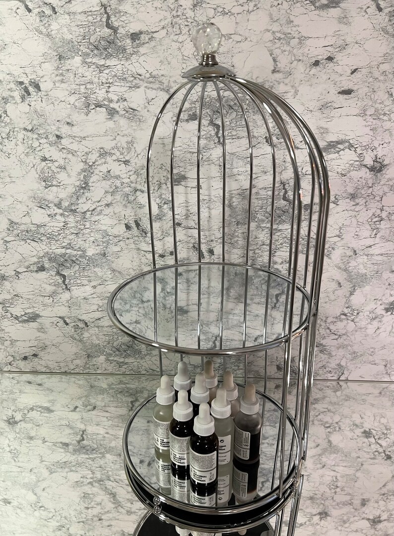 May include: A three-tiered, chrome-finished display stand with mirrored shelves. The top is adorned with a clear, crystal-like sphere. The stand holds several small, clear bottles with white droppers, likely skincare products. The background is a marbled wall.