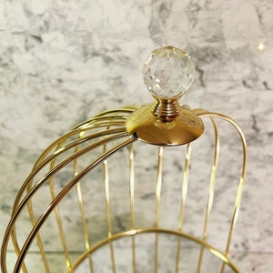 May include: A gold-colored, wireframe basket with a clear, faceted crystal ball on top. The basket has a rounded shape with vertical bars and a decorative, gold-colored base. The background is a light gray and white marble pattern.