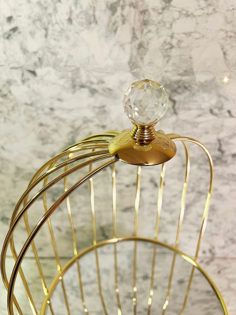 May include: A gold-colored, wireframe basket with a clear, faceted crystal ball on top. The basket has a rounded shape with vertical bars and a decorative, gold-colored base. The background is a light gray and white marble pattern.
