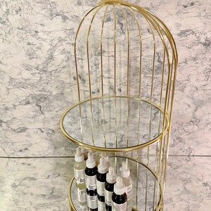 May include: A gold-colored, three-tiered display stand with mirrored shelves. The stand has a cage-like design, topped with a crystal-like sphere. Several bottles with white and black labels are displayed on the shelves.