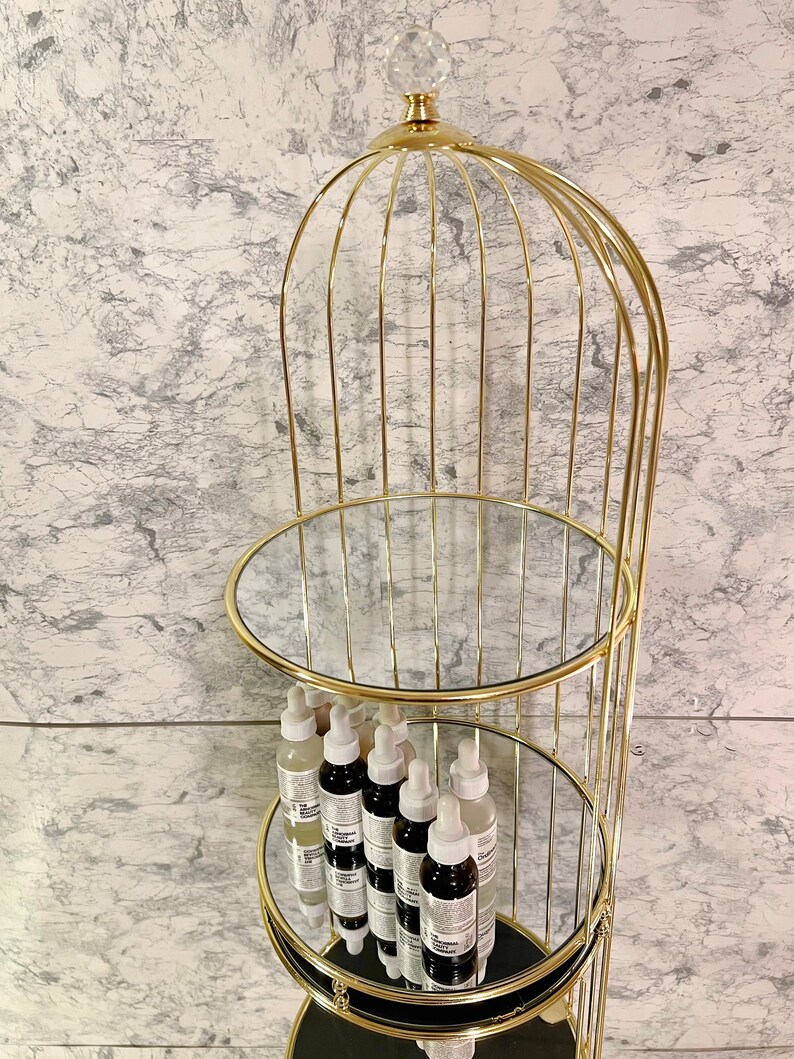 May include: A gold-colored, three-tiered display stand with mirrored shelves. The stand has a cage-like design, topped with a crystal-like sphere. Several bottles with white and black labels are displayed on the shelves.