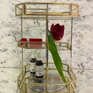 May include: Gold metal tiered stand with three shelves, featuring a red tulip, two clear glass bottles with labels, and a red box. The stand has a decorative design with a mirrored base.