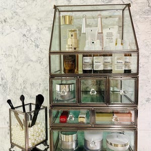 May include: A glass and metal makeup organizer with multiple drawers and a top shelf, filled with various cosmetic products. The organizer has a mirrored base and is accented with crystal knobs and decorative trim. A separate glass container holds makeup brushes.