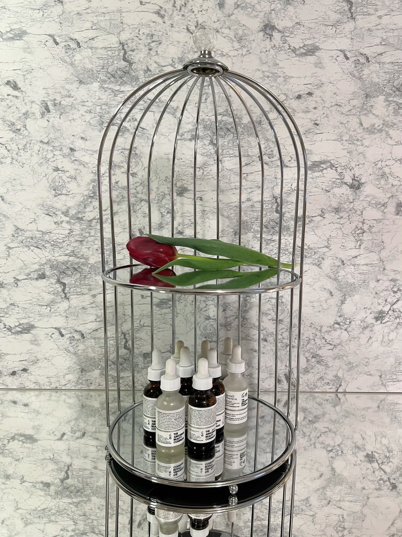 May include: A chrome metal decorative shelf in the shape of a birdcage, with two mirrored shelves. A red tulip and several skincare product bottles are displayed on the shelves. The background is a marble-patterned wall.
