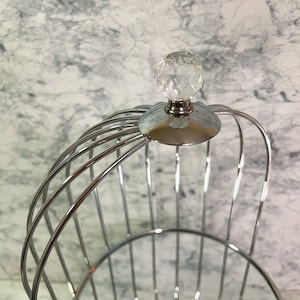 May include: A chrome-finished metal fruit basket with a cage-like design. The basket features a clear, faceted crystal ball on top. The basket is designed with curved metal bars and a circular base, suitable for displaying fruit.