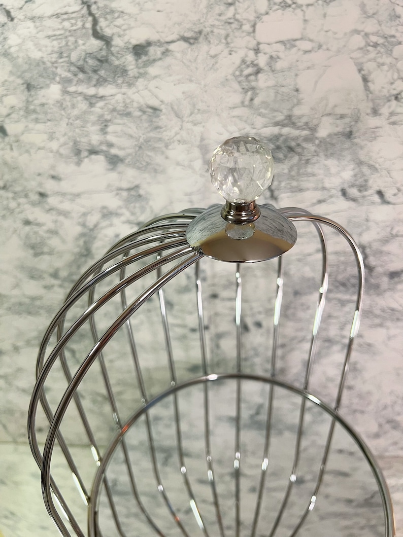 May include: A chrome-finished metal fruit basket with a cage-like design. The basket features a clear, faceted crystal ball on top. The basket is designed with curved metal bars and a circular base, suitable for displaying fruit.