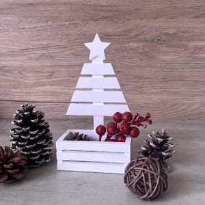 Könnte beinhalten: Eine weiße Weihnachtsbaum-Dekoration aus Holz mit einem Stern oben. Der Baum hat eine kleine Kiste an der Basis, gefüllt mit Tannenzapfen und roten Beerenzweigen. Zwei Tannenzapfen und ein geflochtener Ball sind ebenfalls vorhanden.