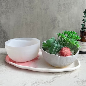 May include: Two white ceramic planters with a textured, irregular rim. The planter on the left is empty, while the planter on the right is filled with small green succulents and a pink flower. The planters are sitting on a white oval tray with a pink, textured surface.