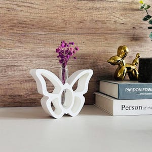 Könnte beinhalten: Weiße Keramik-Schmetterlingsvase mit einem durchsichtigen Glasrohr für Blumen. Die Vase steht auf einem Tisch mit einem Buch und einem goldenen Ballonhund.