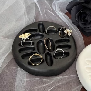 Può includere: Un porta anelli rotondo nero con più scomparti per riporre gli anelli. Il porta anelli è rotondo e ha una finitura liscia e opaca. Diversi anelli sono esposti negli scomparti, tra cui un anello a farfalla, un anello a fascia attorcigliata e un anello a fascia semplice.