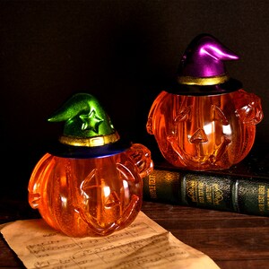 Peut inclure: Deux décorations en forme de citrouille orange translucide avec des visages sculptés, chacune surmontée d'un chapeau de sorcière. L'un est vert avec une étoile, l'autre est violet. Sur une surface en bois sombre, thème Halloween.
