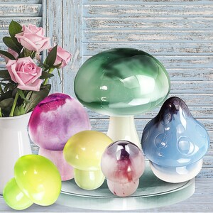 May include: A collection of five ceramic mushroom-shaped decorative objects. The mushrooms are in various colors, including green, purple, yellow, and blue. The mushrooms are arranged on a white surface.