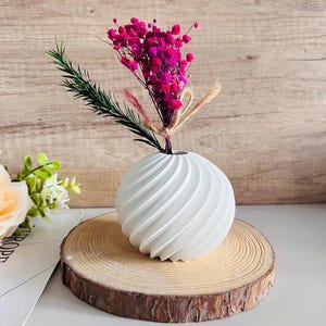 May include: White ceramic vase with a swirl design, holding a bouquet of pink flowers. The vase is sitting on a wooden slice.