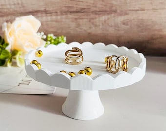 May include: White ceramic cake stand with a scalloped edge. The stand has a small base and holds two gold rings and gold beads.