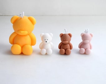 May include: Four small teddy bear candles in different colors: yellow, white, brown, and pink. The candles are all sitting on a white surface.