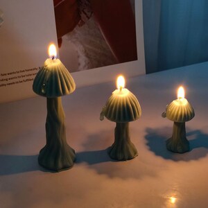 K&ouml;nnte beinhalten: Drei tealblaue kerzen in Pilzform mit einer Flamme, die auf jeder Kerze brennt. Die Kerzen stehen auf einer wei&szlig;en Oberfl&auml;che.