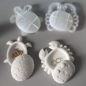 May include: Two translucent silicone molds shaped like a turtle and a crab, alongside two finished white resin trinket dishes in the same animal shapes. The turtle dish holds a gold chain, and the crab dish contains two gold rings.