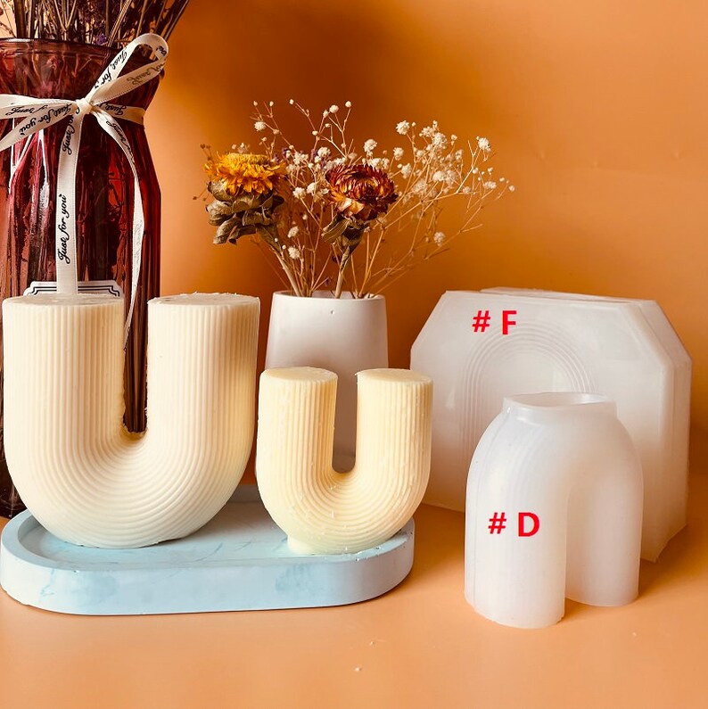 May include: Two white silicone molds for making candles in the shape of the letter U. The molds are on a light blue surface. The mold on the left is larger than the mold on the right. The mold on the right is labeled #D. The mold on the left is not labeled. There is a white silicone mold in the background labeled #F.
