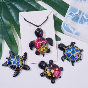 Pode incluir: Uma coleção de pingentes e berloques em forma de tartaruga. Os pingentes apresentam corpos pretos com conchas coloridas cheias de glitter em rosa, azul e dourado. Um pingente está pendurado em um cordão preto. Um molde de tartaruga transparente também é visível.