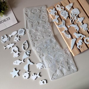 Könnte beinhalten: Ein Set aus weißen Harzformen zum Basteln. Die Formen zeigen verschiedene Meereslebewesen, darunter Fische, Muscheln, Seesterne und eine Qualle. Die Formen sind auf einer Holzoberfläche angeordnet.