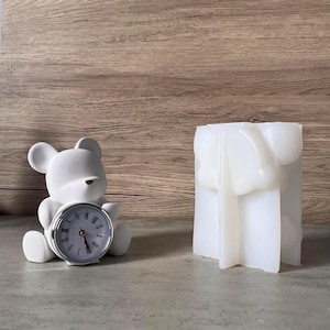 Puede incluir: Un despertador blanco con forma de oso con una esfera plateada y números romanos negros. Al lado, un portavelas rectangular blanco y abstracto. Los objetos están sobre una superficie gris con fondo de vetas de madera.