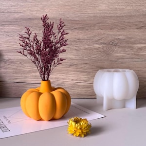 Puede incluir: Un jarrón naranja con forma de calabaza contiene flores moradas secas. Una flor amarilla y un molde blanco con forma de calabaza están cerca. El jarrón está sobre una superficie blanca con un fondo de madera.