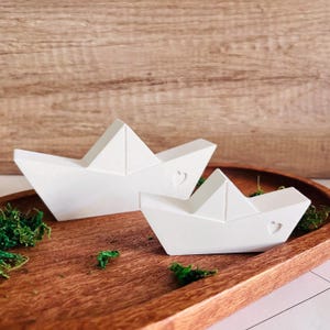 May include: Two white ceramic paper boat sculptures with a heart detail on a wooden tray with green moss.
