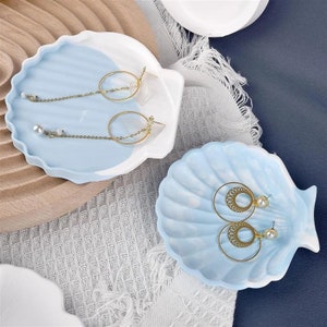 May include: Two blue and white ceramic seashell shaped dishes with gold earrings on top. The dishes are on a white and blue textured surface.