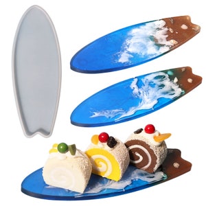 Könnte beinhalten: Drei Miniaturkuchen auf einem blauen und braunen, surfboardförmigen Harztablett. Das Tablett hat ein weißes Wellendesign und braune Akzente. Die Kuchen sind ein gelber Biskuitkuchen mit roten und schwarzen Dekorationen, ein Schokoladenbiskuitkuchen mit weißem Frosting und ein weißer Biskuitkuchen mit grünen Dekorationen.