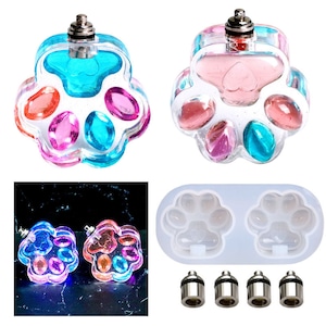 Op de afbeelding: Twee transparante, paw print-vormige harsvormen met kleurrijk hars erin. De vormen zijn gevuld met kleurrijk hars en hebben een klein metalen dopje bovenop. Er is ook een siliconenmal met twee paw print-vormen en vier metalen dopjes.