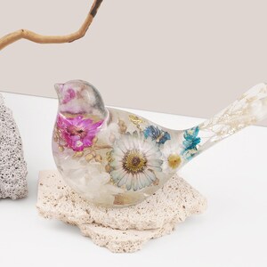 May include: A clear glass bird figurine with dried flowers and leaves embedded inside. The bird is perched on a small, light brown rock.