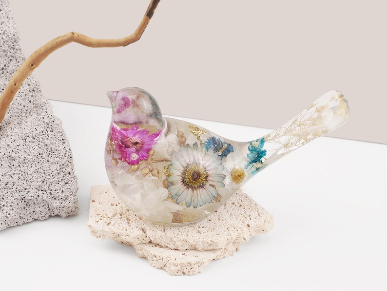 May include: A clear glass bird figurine with dried flowers and leaves embedded inside. The bird is perched on a small, light brown rock.