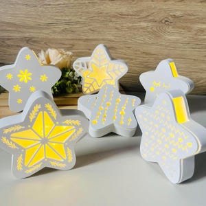May include: A collection of white star-shaped decorative lights with intricate cut-out designs. Each star has a warm yellow glow emanating from within. The stars are arranged on a white surface, suggesting a festive or holiday theme.