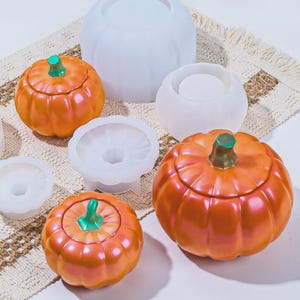 May include: Orange pumpkin-shaped containers with green stems and lids, alongside white silicone molds. The pumpkins have a glossy finish. The items are arranged on a textured, off-white surface, suggesting a fall or Halloween theme.