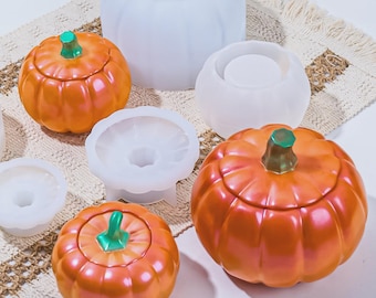May include: Orange pumpkin-shaped containers with green stems and lids, alongside white silicone molds. The pumpkins have a glossy finish. The items are arranged on a textured, off-white surface, suggesting a fall or Halloween theme.