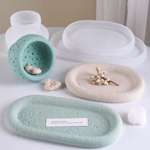 May include: An assortment of decorative trays and containers in various shapes and colors. Includes a light green oval tray with text, a light blue textured bowl, and a white oval tray with a sprig of flowers. Other trays are white and translucent.