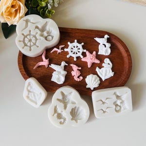 Könnte beinhalten: Eine Sammlung weißer und rosa Silikonformen mit nautischem Thema, darunter ein Steuerrad, Seesterne, Seepferdchen, Möwen und Muscheln. Die Formen sind auf einem Holztablett angeordnet, ideal zum Basteln oder Backen.