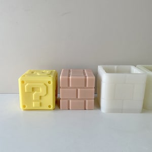 Puede incluir: Tres bloques impresos en 3D de diferentes colores inspirados en el videojuego Super Mario Bros. Los bloques son amarillos con un signo de interrogación, rosa con un patrón de ladrillos y blanco con un patrón de ladrillos.