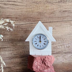 Könnte beinhalten: Weiße, hausförmige Uhr mit silbernem Zifferblatt und schwarzen römischen Ziffern. Die Uhr steht auf einem roten Steinsockel.
