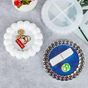 May include: Three round silicone moulds for resin crafts. The moulds are white and have a raised, textured edge. One mould has a divided interior, another has a smooth interior, and the third has a blue interior with a white rectangle and a green mushroom.