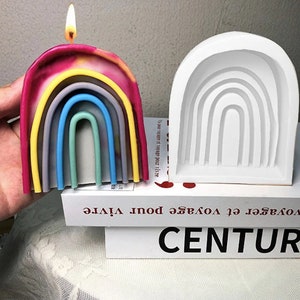 May include: A white silicone mold shaped like a rainbow with seven arches. A rainbow candle is next to the mold. The candle is pink, yellow, gray, blue, green, and yellow. The candle is lit.