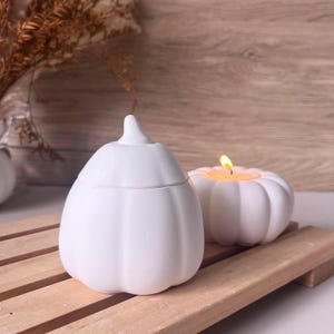 Puede incluir: Contenedor blanco de cerámica con forma de calabaza con tapa y una pequeña vela blanca con forma de calabaza con una llama encendida. Los objetos están sobre una superficie de madera, con un fondo de madera.