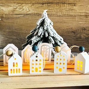 May include: A string of white ceramic houses with light bulbs inside, decorated with small wooden beads and a green artificial Christmas tree in the background.