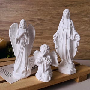 Könnte beinhalten: Drei weiße Engelsstatuen in verschiedenen Größen. Die größte Statue zeigt eine Frau mit ausgestreckten Armen. Die anderen beiden Statuen sind Engel mit Flügeln, einer kniend und der andere stehend mit gefalteten Händen.