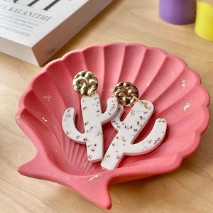 May include: A pair of white cactus-shaped earrings with gold accents. The earrings are displayed on a pink seashell-shaped dish.