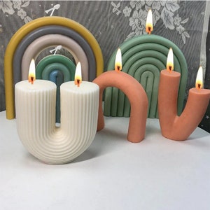 May include: Three colorful candles shaped like rainbows and abstract arches. The candles are lit and have a white, orange, and green color scheme.