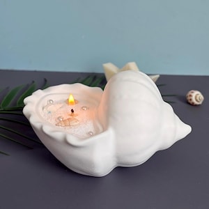 Puede incluir: Un portavelas de cerámica blanca con forma de concha marina que contiene cera blanca, una mecha encendida y elementos decorativos incrustados, incluyendo pequeñas conchas y perlas blancas. El recipiente tiene un acabado mate y texturizado.