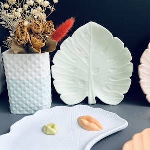 May include: Three ceramic dishes in different shapes and colors. The first dish is a white leaf-shaped dish. The second dish is a white fan-shaped dish with two small colorful candies on it. The third dish is a pink round dish.