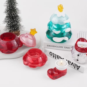 May include: A set of five Christmas-themed candle holders made of glass. The candle holders are shaped like a Christmas tree, a snowman, a boot, and two round bowls. The candle holders are all red and clear glass. The candle holders are sitting on a white surface.