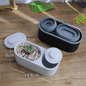 May include: Two oval-shaped planters, one white and one black, with gray interior components. The white planter, measuring 19.8 cm long and 8 cm high, contains a succulent and decorative stones. The black planter has two gray inserts. Lids are included.